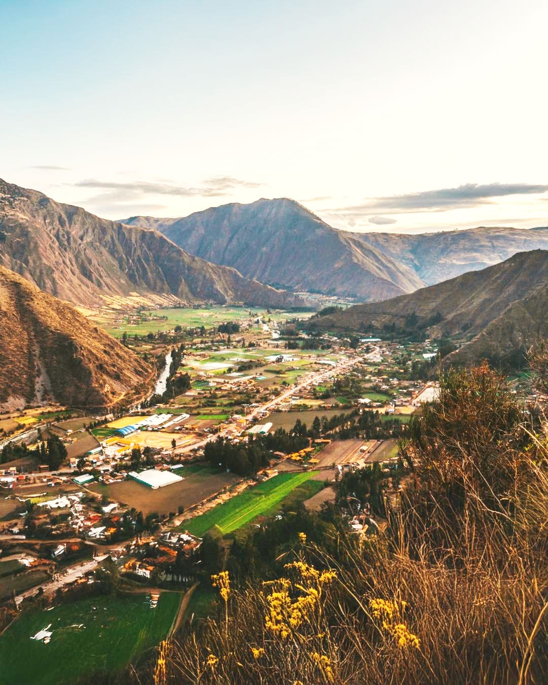 An aerial view of the Sacred Valley in Peru, showcasing lush farmlands, a winding river, and surrounding mountains under a serene sky.