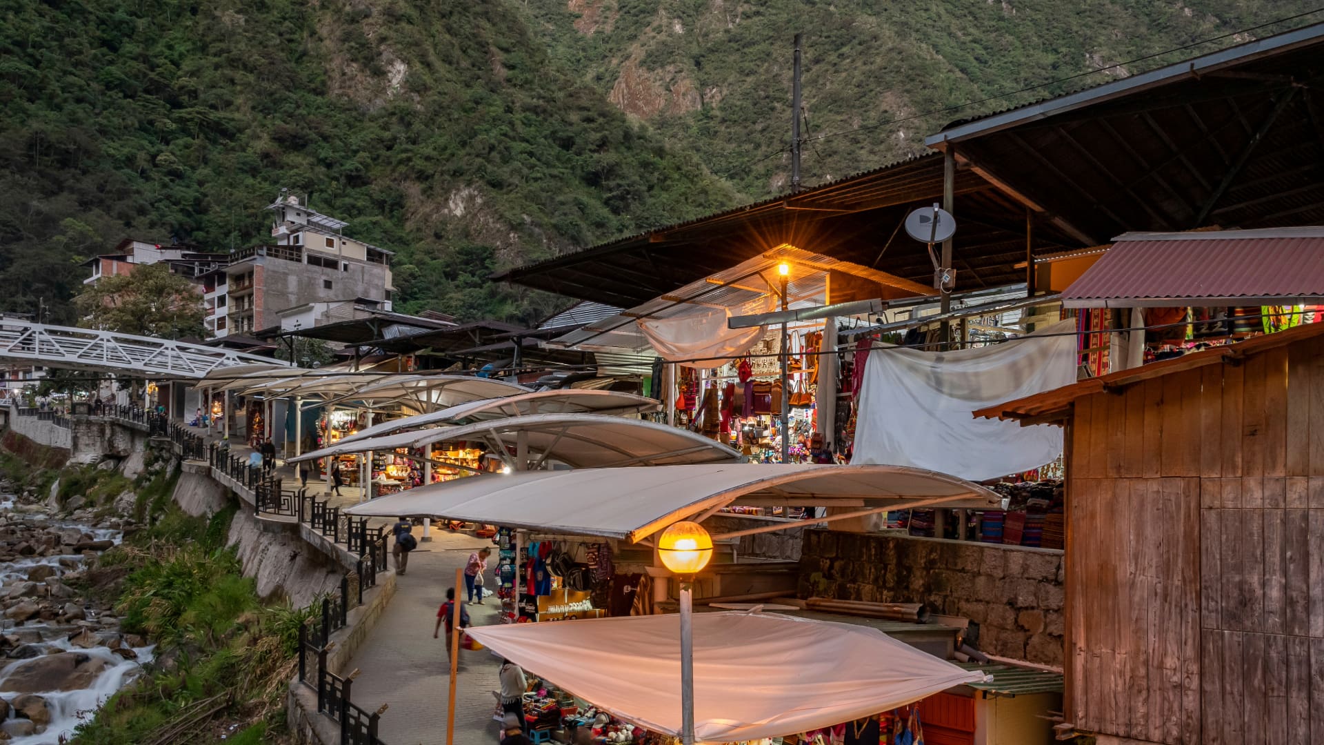 Evening view of the bustling artisan market in Aguas Calientes, Peru, with vibrant stalls, colorful crafts, and a scenic backdrop of lush green mountains.