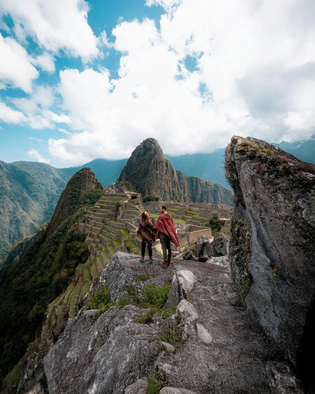 A couple dressed in traditional Andean ponchos enjoying a breathtaking view of Machu Picchu from a high vantage point, with terraces and Huayna Picchu in the background under a blue sky.