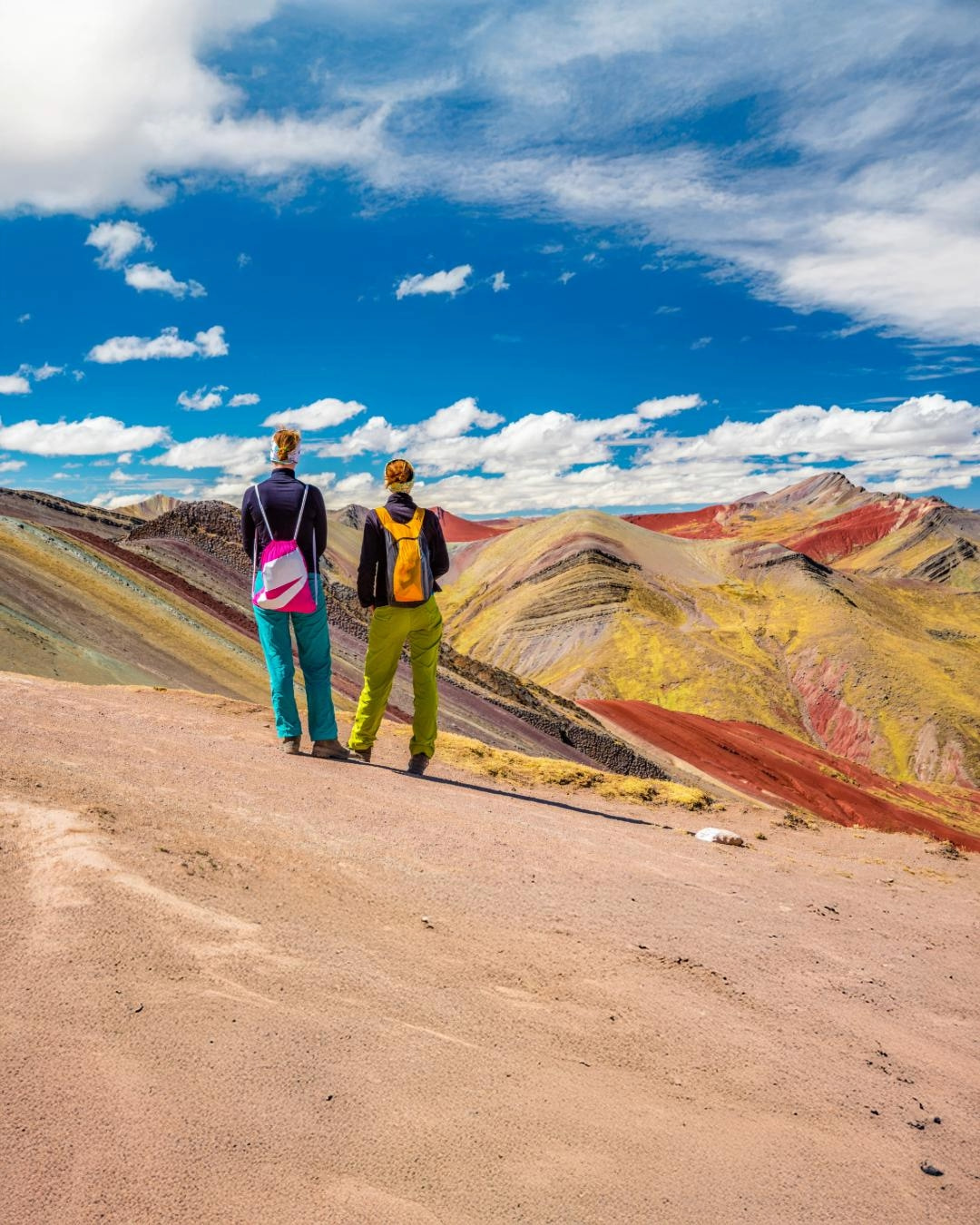 Two hikers standing on a sandy plateau, overlooking the colorful Rainbow Mountain in Peru under a bright blue sky.