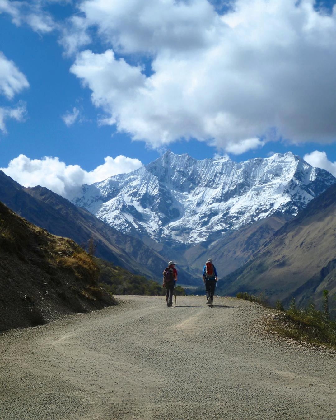 Two hikers with backpacks on a trail leading towards a snow-capped mountain range on a sunny day.