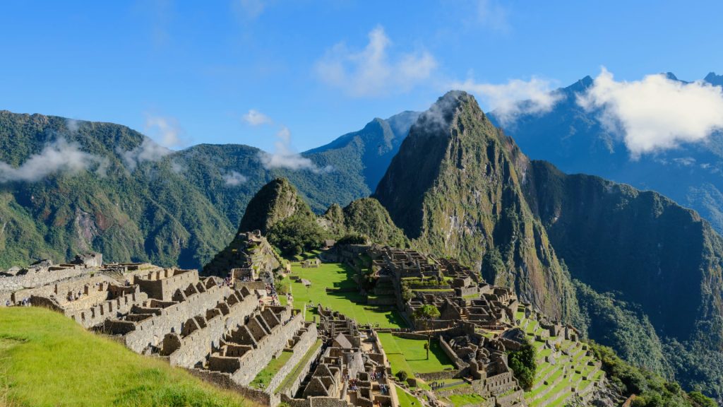 Panoramic view of Machu Picchu ruins surrounded by lush green mountains and the towering Huayna Picchu mountain, under a clear blue sky.