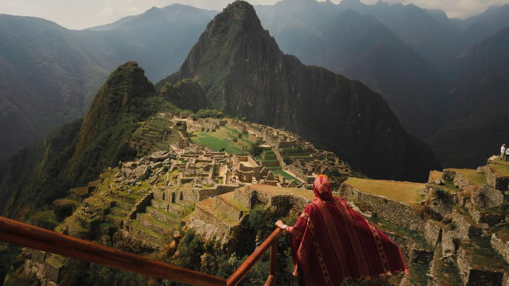 Persona con poncho andino observando Machu Picchu desde un mirador, con terrazas verdes, construcciones incas y Huayna Picchu de fondo-Xplora Machu Picchu