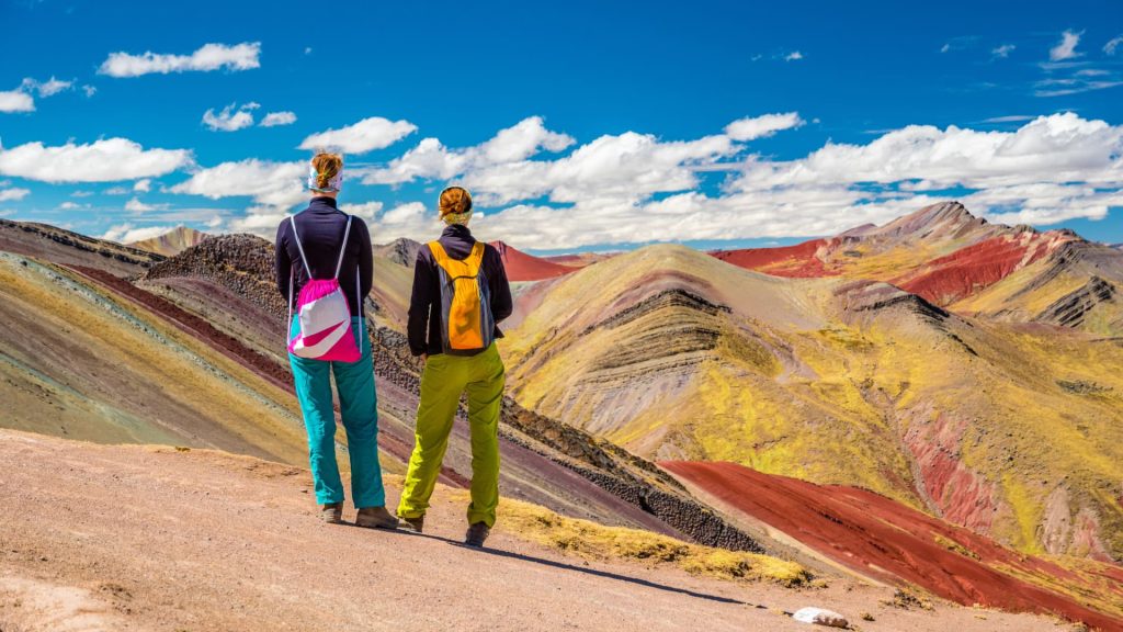 Two hikers standing on a viewpoint, admiring the colorful mountains of Palccoyo under a bright blue sky - Xplor Machupicchu