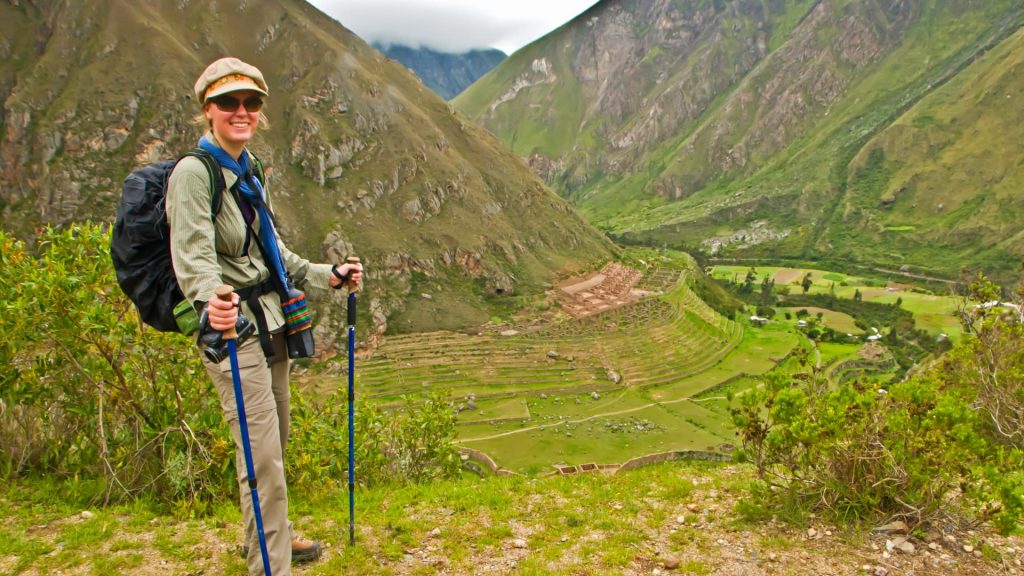 A smiling hiker with trekking poles and a backpack on the Inca Trail, overlooking ancient Inca terraces and stunning Andean landscapes.