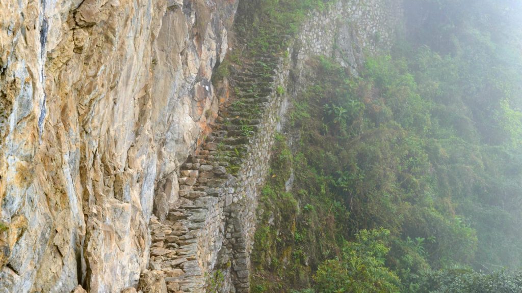 The Inca Bridge at Machu Picchu, an ancient stone pathway built along a steep cliff, partially covered by mist and surrounded by lush vegetation.