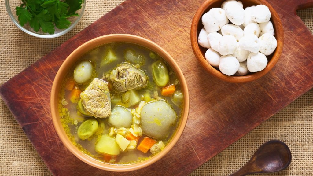 Traditional Andean caldo de mote, a rich soup with beef, potatoes, corn, and vegetables, served with chulpi corn on a wooden board – Xplor MachuPicchu