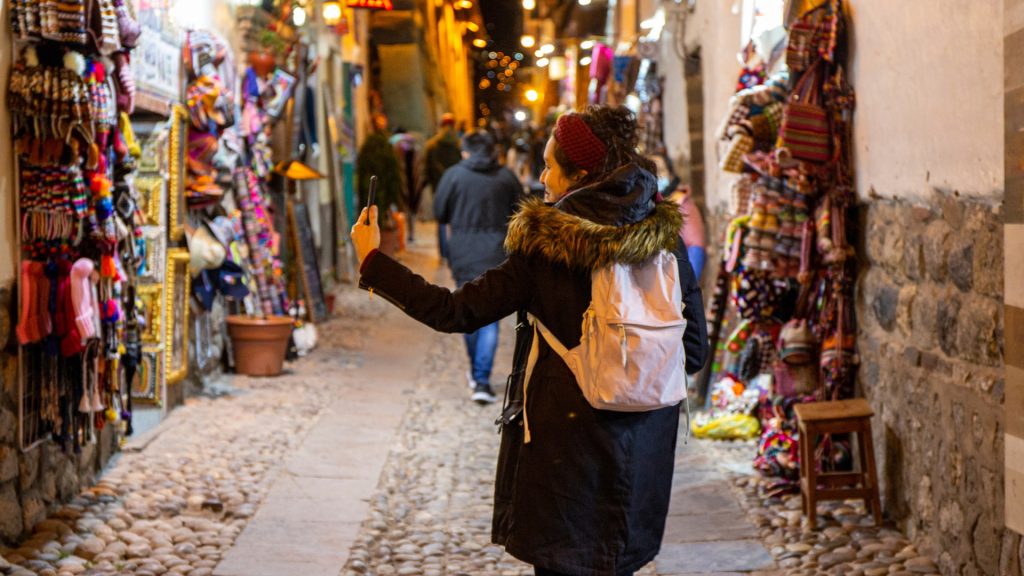Woman walking along a cobbled street lined with colorful handicraft stores, experiencing the cultural charm of Cusco’s nightlife - Xplor Machupicchu