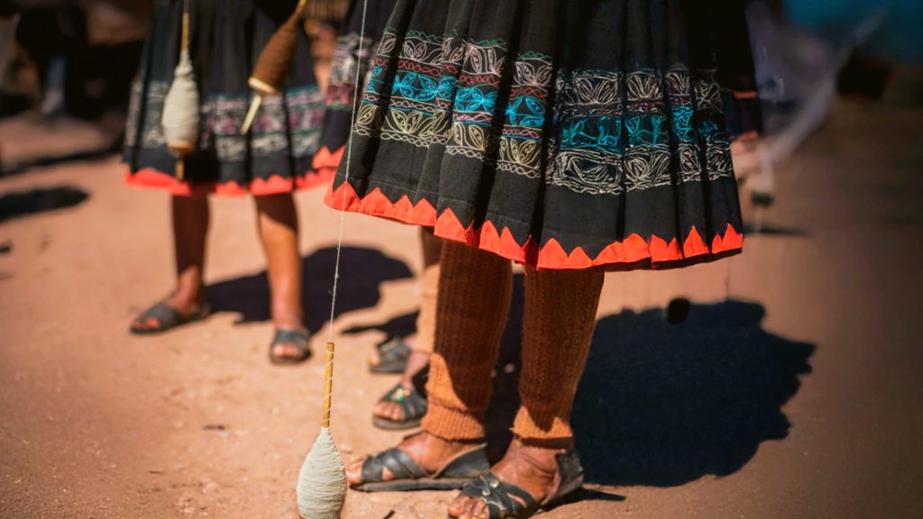 Traditional Andean women spinning wool using ancient techniques, wearing embroidered skirts with vibrant designs - Xplor Machu Picchu.
